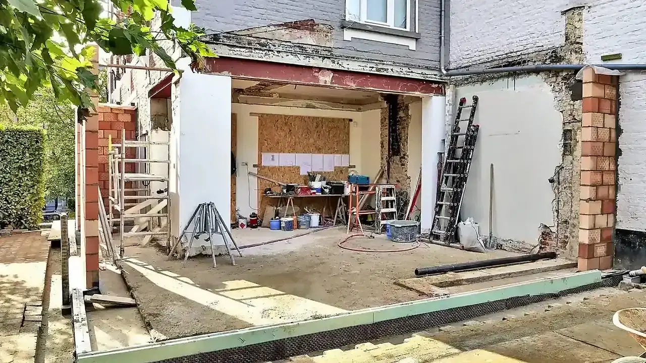 Une extension de maison en cours, avec un mur arrière ouvert, un échafaudage, une échelle, un établi et des matériaux dans la cour